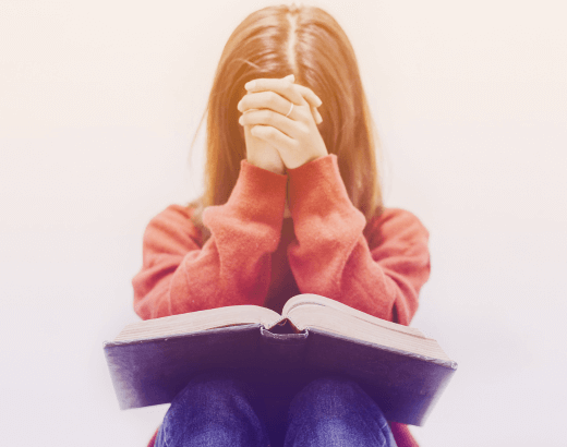 mulher orando com uma Bíblia da Mulher em seu colo. ela veste um casaco de magas longas vermelho, calça jeans e está com as mãos juntas em frente ao rosto em oração