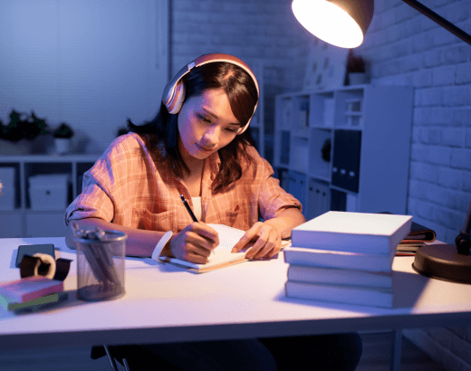 garota estudando com caderono apiado sobre uma mesa onde tem livros, luminária de mesa, canetas e usa um fone de ouvido (1)