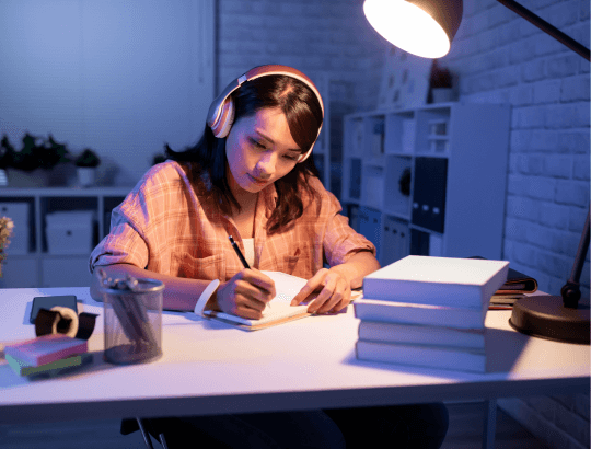 garota estudando com caderono apiado sobre uma mesa onde tem livros, luminária de mesa, canetas e usa um fone de ouvido (1)